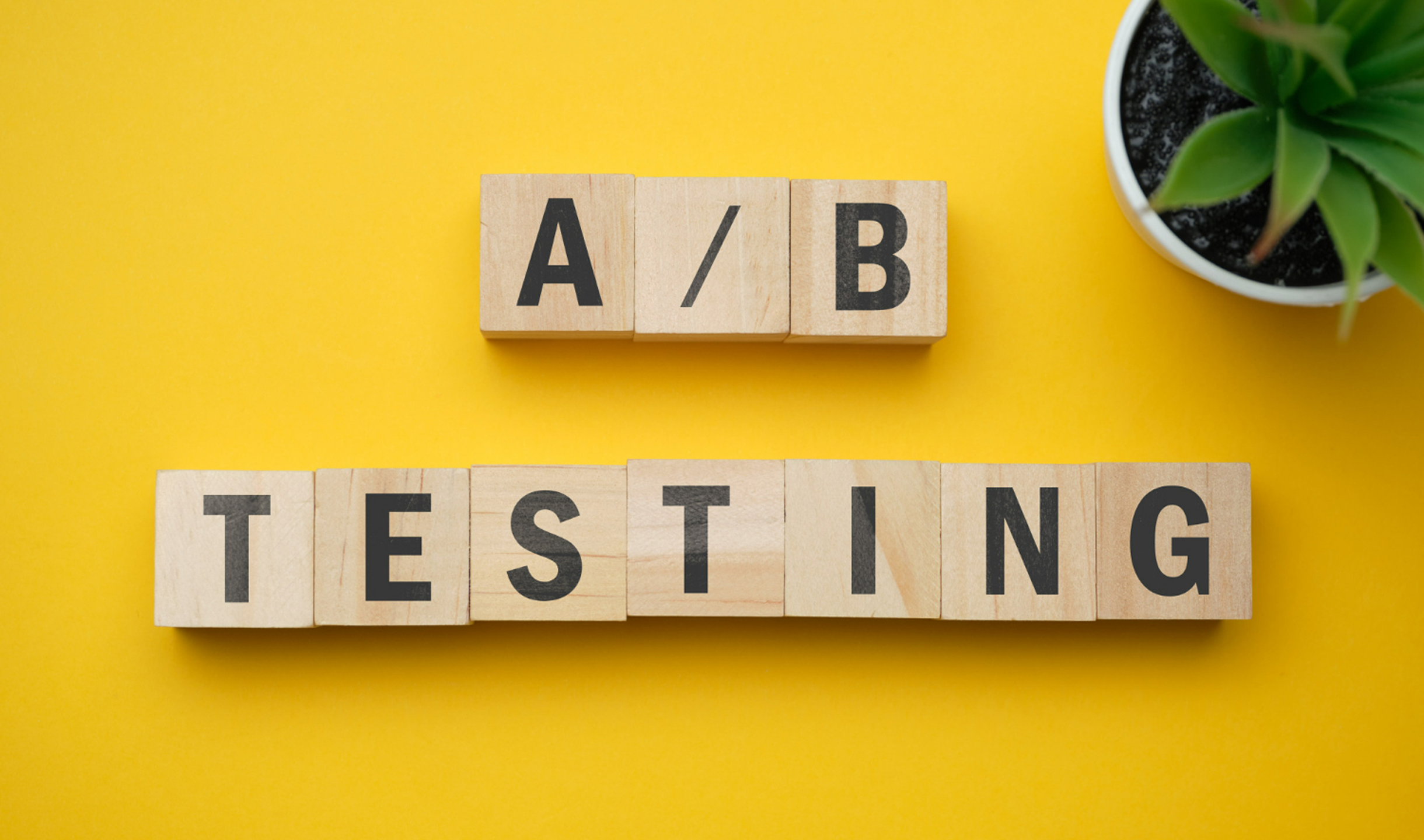 A/B Testing이라는 단어가 우드 블록 위에 적혀 있다. 주위 배경은 노란색이고 오른쪽 상단에 화분이 하나 있다.